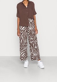 Bruine button-up overhemd met korte mouwen gecombineerd met witte en bruine zebraprint wijde broek. Witte sneakers maken de outfit compleet.