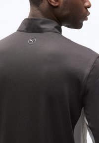 Κοντινό πλάνο ενός ατόμου που φοράει μαύρο πουκάμισο με ψηλό γιακά και μικρό λευκό λογότυπο Puma στο πάνω μέρος της πλάτης.