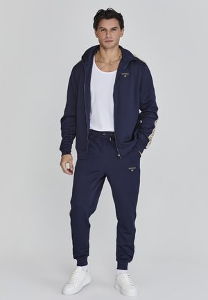 Chándal marino con detalles dorados, que incluye una chaqueta con cremallera y cuello alto, y pantalones de chándal ajustados. Combinado con una camiseta blanca y zapatillas blancas.
