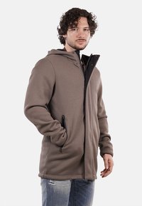 Giacca marrone a coste con zip frontale, cappuccio e due tasche laterali. Il tessuto sembra testurizzato con una vestibilità casual e rilassata.