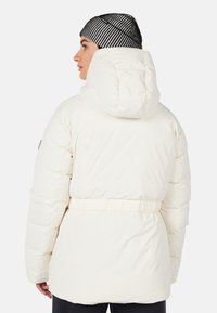 Person med en hvid polstret jakke med hætte og en sort-hvid stribet beanie, vist fra ryggen.