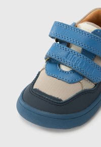 Αθλητικό παπούτσι με αυτοκόλλητα λουράκια, μπλε δερμάτινο και μπεζ υφασμάτινο επάνω μέρος, στρογγυλεμένη μύτη, λαστιχένια σόλα με υφή για καλύτερο κράτημα και ραφές ως διακοσμητικό στοιχείο.