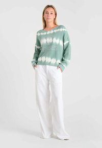 Lichtgroene tie-dye trui met geribde textuur en een losse pasvorm, gecombineerd met witte wijde broekspijpen. De trui heeft bedrukte tekst aan de voorkant.