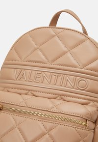 Beige leather backpack with quilted pattern, featuring a prominent embossed logo and a zippered front pocket with gold hardware.