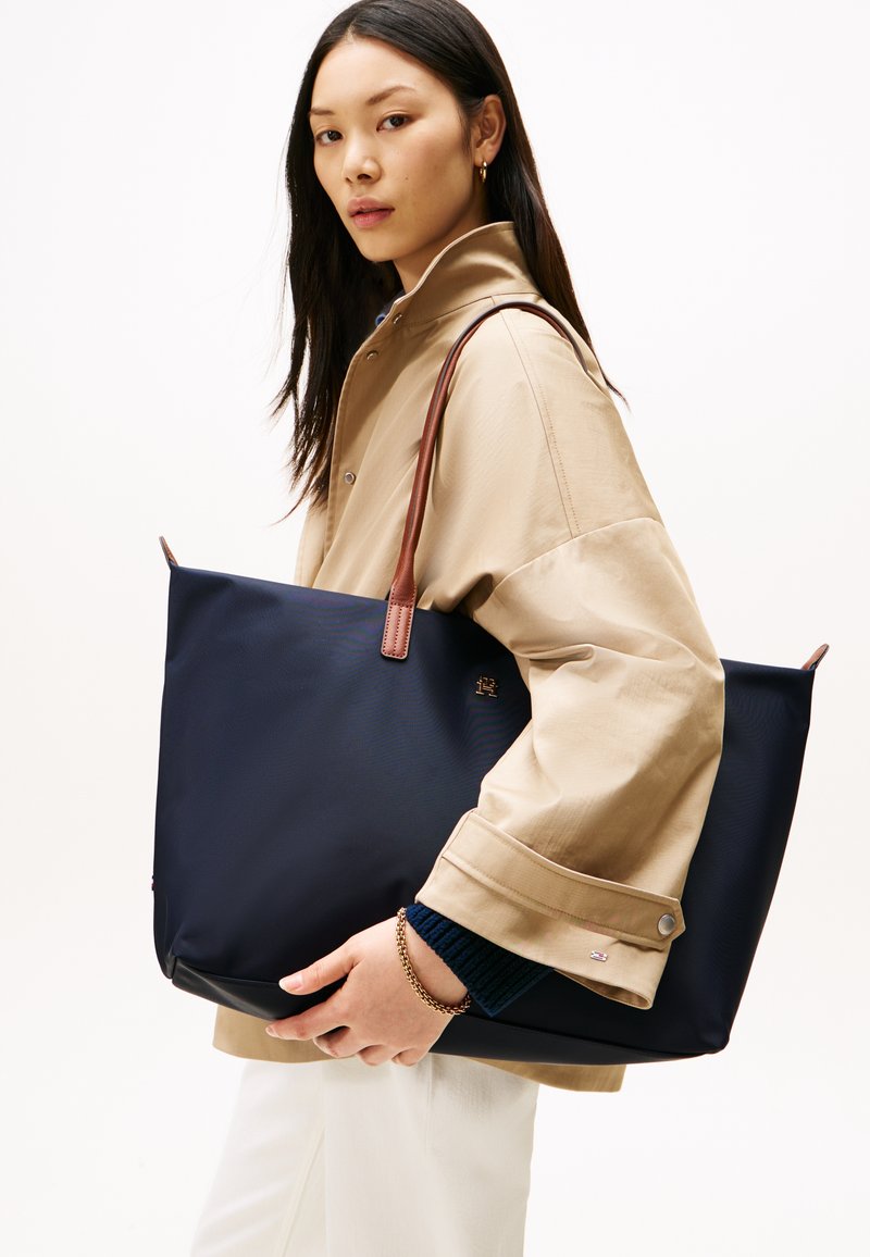 Kvinde iført beige jakke og hvide bukser bærer stor navy-blå taske med brune håndtag på skulderen.