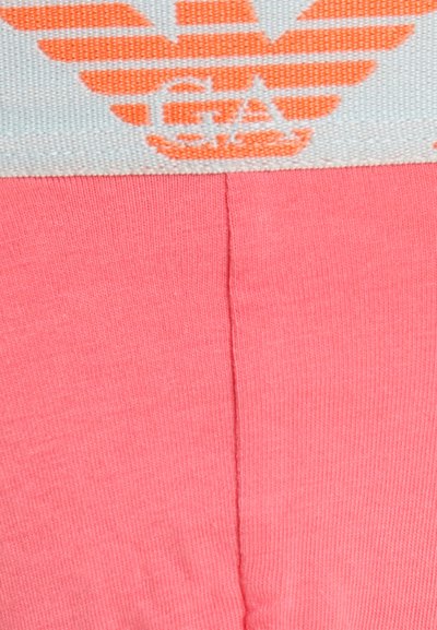 Κοντινό πλάνο σε ροζ ύφασμα με λευκή και πορτοκαλί ζώνη στη μέση που φέρει ένα στυλιζαρισμένο λογότυπο με φτερά και μερικό κείμενο.