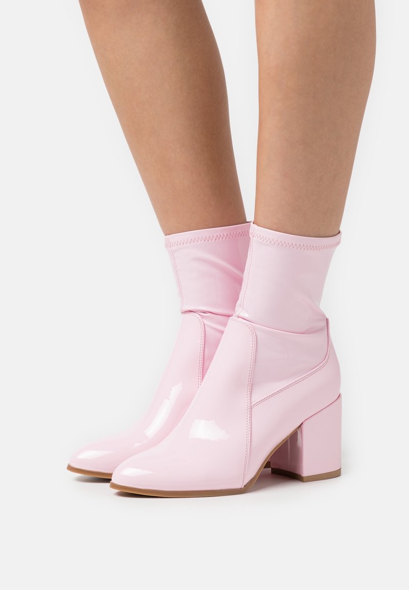 Jambes portant des bottines rose pastel brillantes avec talons carrés et matière supérieure douce et extensible sur un fond uni.