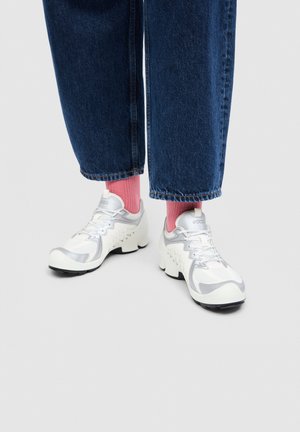 Persona valkā baltus un pelēkus sporta apavus ar rozā zeķēm un zilas džinsa bikses, stāv uz vienkārša gaiši pelēka fona.