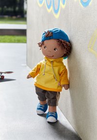 Doll wearing a yellow hoodie, gray pants, and blue shoes, with curly brown hair and a blue cap, standing against a wall.