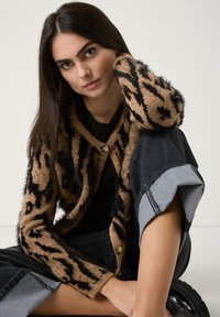 Flauschige Leopardenmuster-Cardigan in Beige und Schwarz mit einer Knopfschließe. Kombiniert mit dunkelblauen Jeans mit hochgekrempelten Säumen.