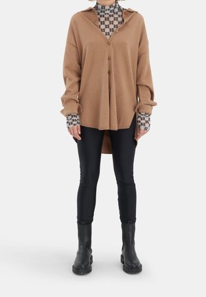 Frau trägt übergroßes, camel-farbenes Hemd mit Knöpfen über gemustertem Rollkragenpullover, dazu schwarze Leggings und schwarze Lederstiefel, steht vor weißem Hintergrund.