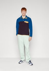 Fleece long-sleeve polo in blue, purple, and mustard with a chest pocket. Paired with light green pants and sneakers featuring a white sole.