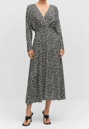 Kobieta ubrana w midi sukienkę w czarno-białe plamki z długim rękawem i dekoltem w serek oraz czarne sandały na obcasie.