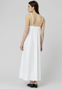 Robe maxi blanche avec des fines bretelles et un dos élastique, présentant une texture lisse et une silhouette fluide, associée à des chaussures plates noires.