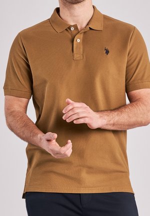 Uomo che indossa una polo marrone a maniche corte con due bottoni, tiene le mani davanti, con un piccolo logo nero ricamato sul petto.