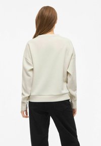 Mujer con cabello castaño claro, vestida con un sudadera de manga larga color blanco roto y pantalones negros, de espaldas contra un fondo blanco plain.