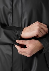 Chaqueta negra impermeable con una textura suave, que presenta un puño abotonado y un discreto detalle en las costuras. Primer plano de manos ajustando la manga.