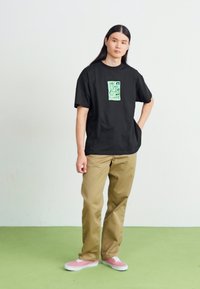 T-shirt en coton noir avec un graphique vert sur la poitrine, associé à un pantalon beige et des baskets roses, se tenant sur un sol vert.