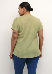 Olive zelený top s krátkým rukávem a texturovaným mřížkovým vzorem, mírně zakřivený lem, nošený s modrými skinny džínami. Zadní pohled ukazuje tetování na ruce.