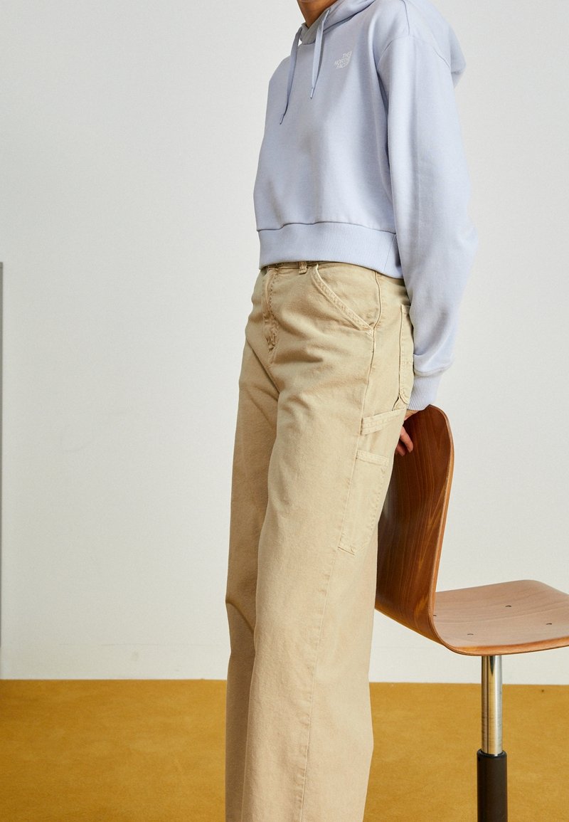 Sweat à capuche court bleu clair avec cordons de serrage, associé à un pantalon cargo beige à jambes larges avec poches latérales, présenté à côté d'une chaise en bois.