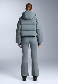 Grå pufferjacka med huva och voluminös design, kombinerad med åtsittande, blixtlåsförsedda grå byxor. Slät yta och modern silhuett.