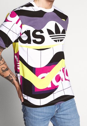Homme portant un t-shirt Adidas coloré avec des motifs abstraits et des logos en noir, violet, jaune et rose, associé à un jean bleu.