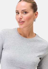 Hellgraues Langarmshirt mit Rundhalsausschnitt, aus weichem Stoff. Nahaufnahme eines Modells mit silbernen Creolen.