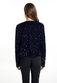 Veste bleu marine à sequins avec une surface texturée, encolure ronde, manches longues et coupe droite, associée à un pantalon noir.