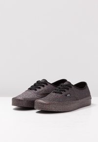 Svarta sneakers med en glittrig finish, som har en rund tå, svarta snören och platta gummisulor. Över ytan har de en strukturerad glittrande effekt.