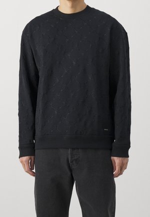 Uomo che indossa un maglione nero a maniche lunghe con un delicato motivo a diamante testurizzato e pantaloni neri, in piedi contro uno sfondo semplice.