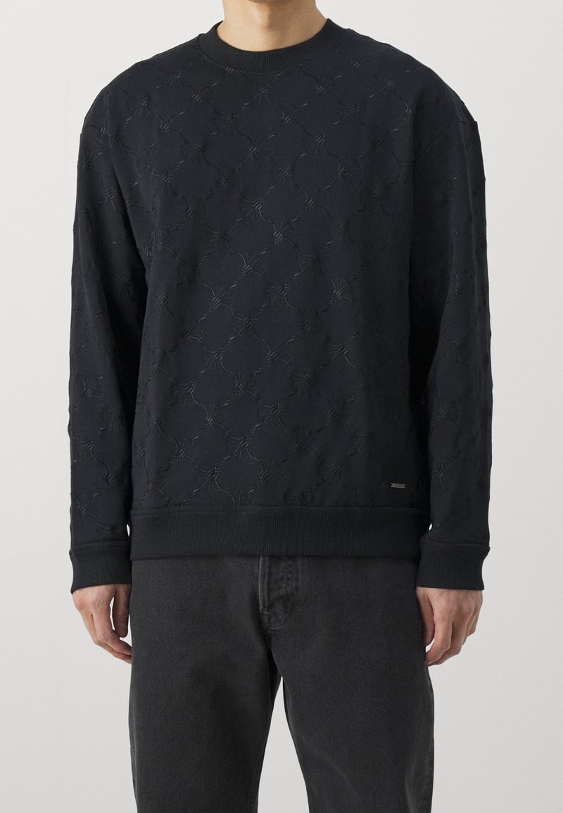 Homme portant un pull noir à manches longues avec un subtil motif losange texturé et un pantalon noir, debout devant un fond uni.