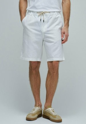 Hombre con pantalones cortos blancos de algodón con cordón y bolsillos y zapatillas beige, de pie sobre un fondo gris neutro.