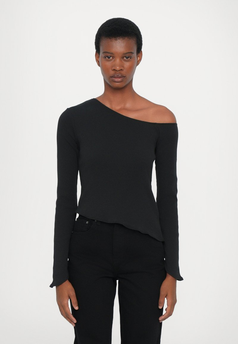 Top nero asimmetrico a maniche lunghe con una spalla scoperta, caratterizzato da un design aderente e una texture morbida, abbinato a jeans neri.