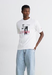 Vit bomull T-shirt med röd och svart grafisk tryck, med texten "23" och "JORDAN", rund hals, korta ärmar, avslappnad passform. Ljust denimbyxor.