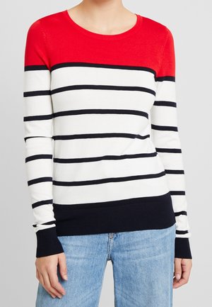 Kvinde iført en langærmet sweater med rød bærestykke, hvid krop med sorte vandrette striber og mørk kant, kombineret med lyseblå jeans.