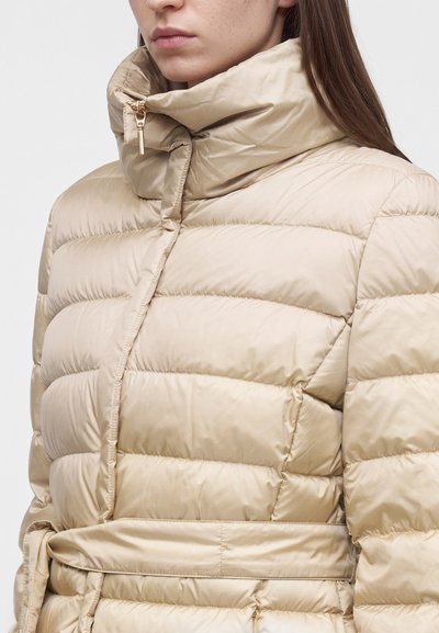Femme portant une doudoune beige avec col montant, fermeture éclair et ceinture, montrée des épaules à la taille sur un fond uni.