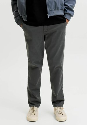 Personne portant un pantalon gris foncé, des baskets beiges, une chemise noire et une veste bleue avec les mains dans les poches.