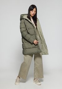 Olivgrön pufferjacka med huva, med ett quiltat mönster, två fickor och ett lätt foder invändigt. Kombineras med beige vida byxor och sneakers.