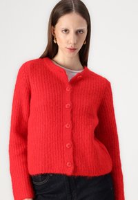 Cardigan rouge tricoté avec un motif texturé, sept boutons roses et un col en V. Matière douce avec des manches longues et une coupe cropped.