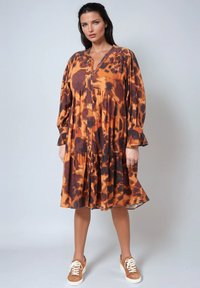 Femme aux cheveux bruns portant une robe à boutons orange et marron à motifs, à manches longues et des baskets, se tenant devant un fond uni.