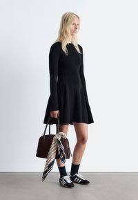 Schwarzes langärmliges Strickkleid mit einem ausgestellten Rock, kombiniert mit einer braunen Tasche, die einen Schal enthält. Schwarze und weiße Sneakers und dunkle Socken vervollständigen den Look.