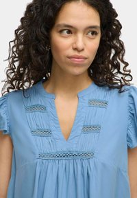 Light blue tunic top with a V-neckline, pleated front, and lace insets. Features short ruffled sleeves and a loose fit. Soft cotton fabric.