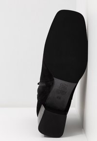 Botte à talon haut en daim noir avec un bout carré et un talon texturé. Dispose d'une fermeture éclair latérale et d'une semelle en caoutchouc lisse et plate. Taille indiquée 4½.