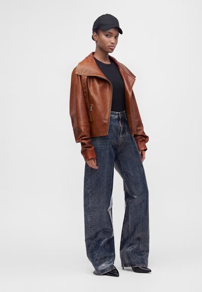 Femme portant une veste en cuir marron, un t-shirt noir, un jean large et foncé, des chaussures noires pointues et une casquette de baseball noire sur un fond uni.