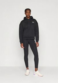 Camisola com capuz preta com um bolso frontal e cordão, combinada com leggings pretas justas, usada com sapatos desportivos de cor clara.