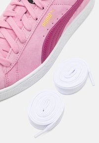 Baskets en daim rose avec un accent rose foncé et une semelle en caoutchouc blanche. Comprend deux lacets blancs soigneusement enroulés à côté de la chaussure.