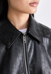 Svart skinnjakke med stor krage, sølvfarget glidelås og CK-logo preget på stoffet. Jevn tekstur med en lett glans.