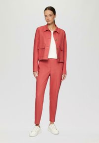Korallefarbener Blazer mit zwei Fronttaschen und goldenen Knöpfen, kombiniert mit passenden schmalen Hosen und weißen Turnschuhen. Glatte Stoffstruktur.