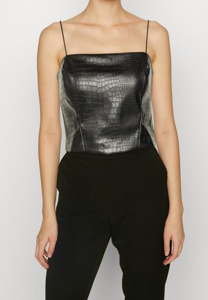 Femme portant un haut sans manches en simili cuir noir à texture crocodile avec fines bretelles et un pantalon noir, debout devant un fond uni.