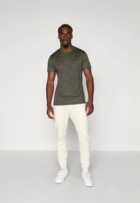 Olivengrøn t-shirt med en glat tekstur, kombineret med hvide tætsiddende joggingbukser. Modellen står med armene afslappede, og hvide sneakers fuldender looket.